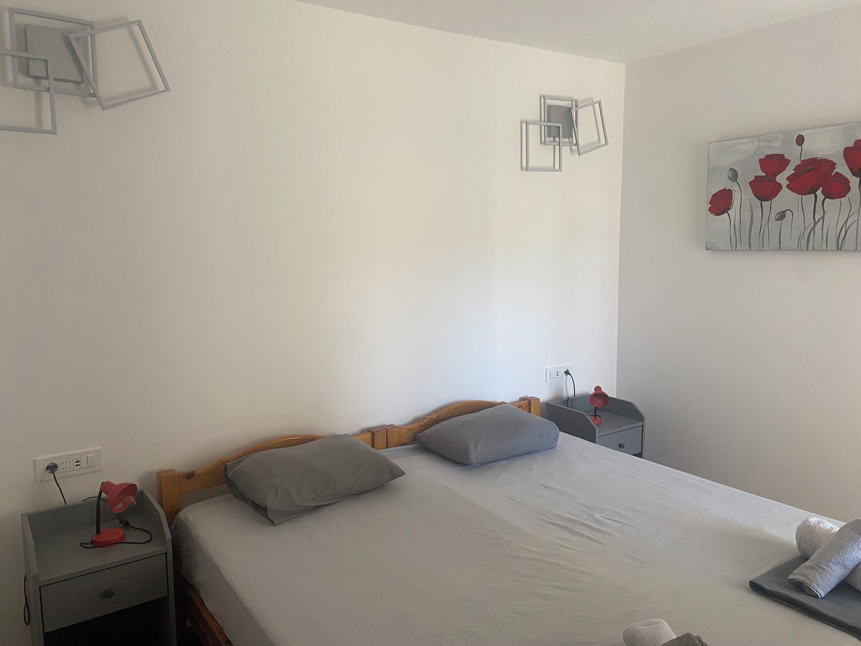 A neatly made bed with grey and white bedding, a wooden headboard, and a grey nightstand with a red lamp.