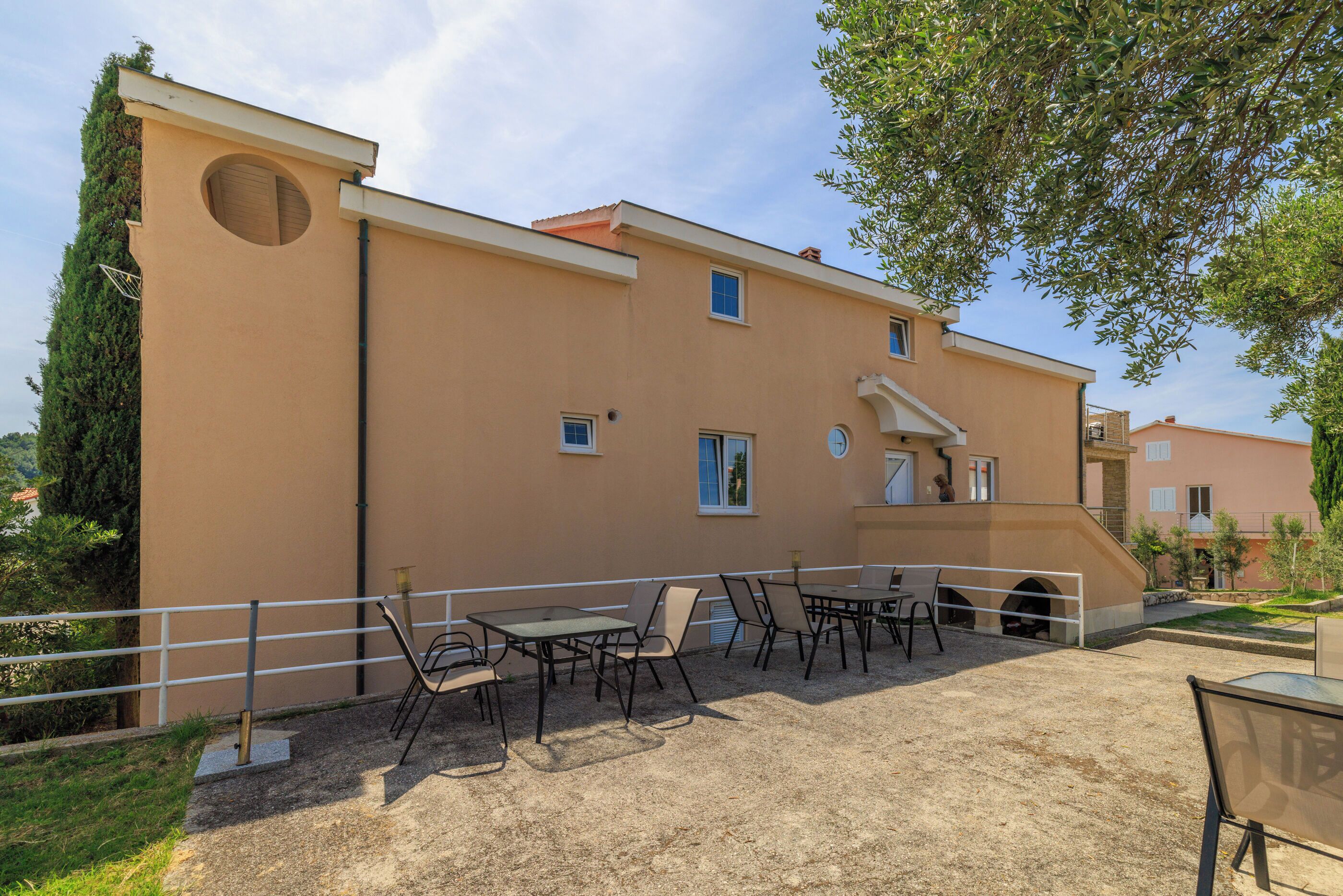A building with a flat roof, a circular window, and a small door. There are outdoor tables and chairs.