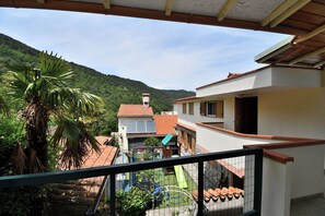 Un barrio residencial con una casa de techo rojo con tejas, un balcón con barandilla de metal y una vista de una colina verde al fondo.
