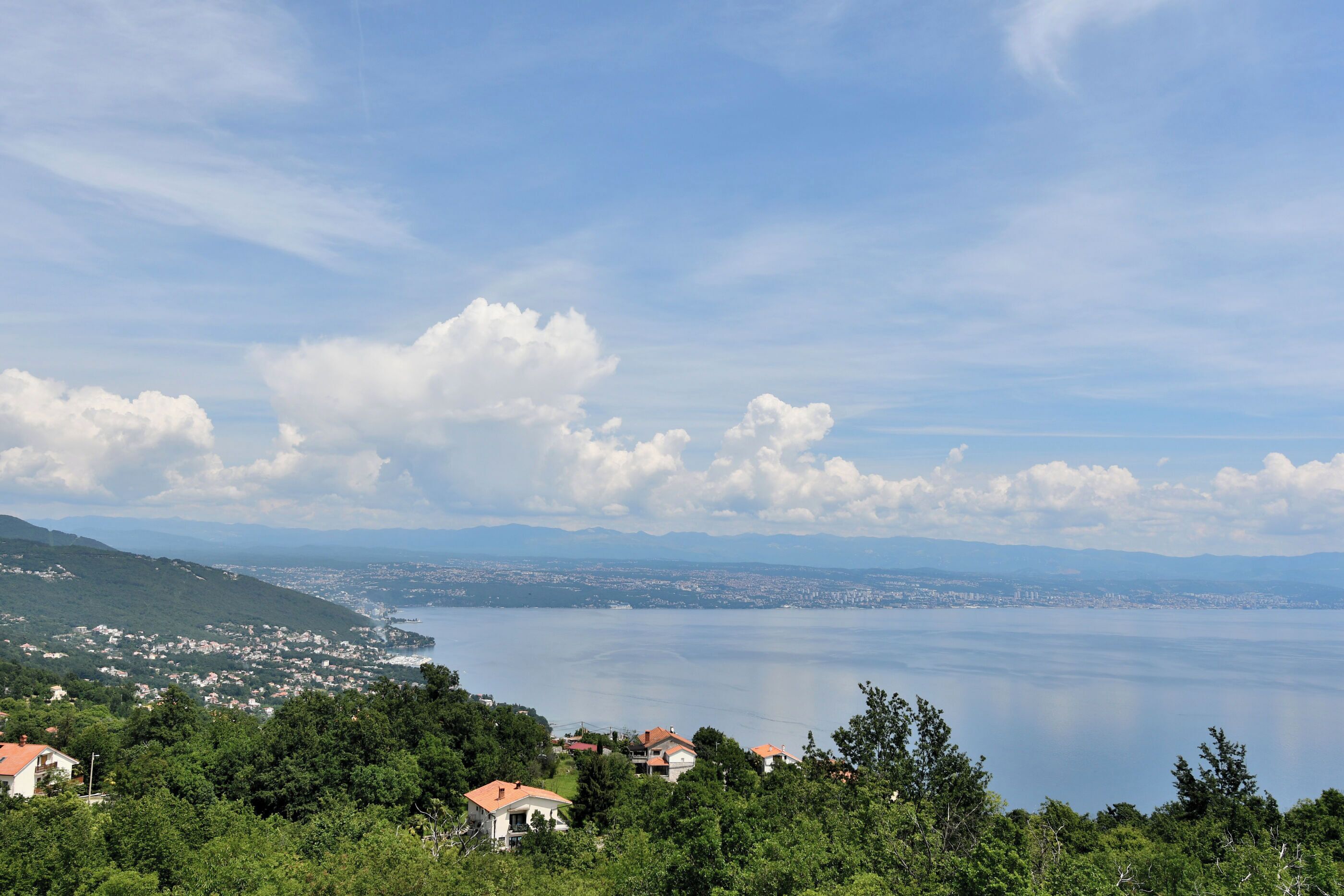 A coastal town with houses on a hillside, overlooking a calm sea and distant mountains.