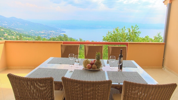 A dining table with wicker chairs and a fruit bowl, overlooking a scenic view of mountains and a body of water. - Apartments Rita (Lovran)