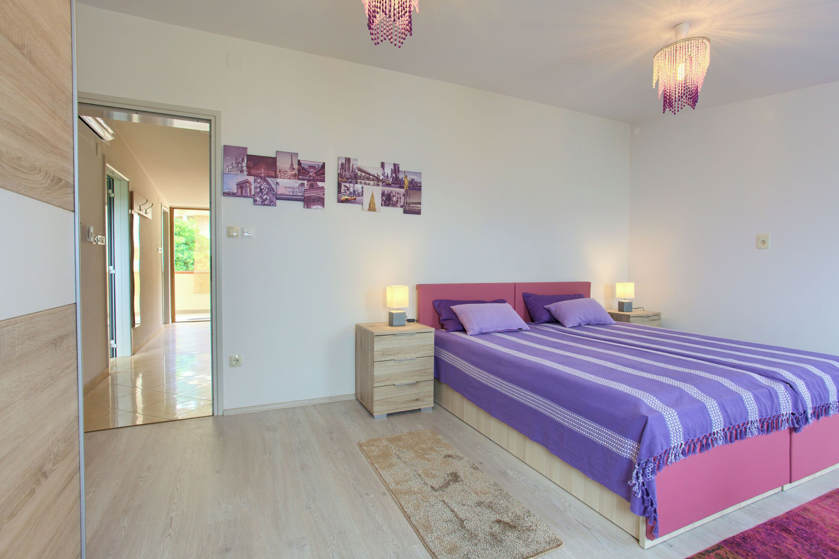 A modern bedroom with a large bed, bedside tables, a chandelier, and a hallway leading to another room.