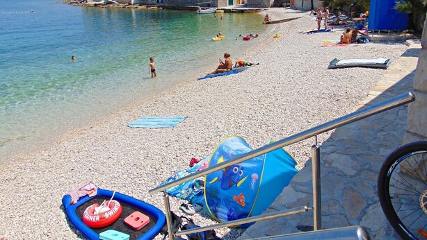 A pebbled beach with clear water, people sunbathing, and a row of stone buildings in the background. - Apartment Fran (Postira)