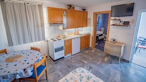 Una cocina compacta con armarios de madera, una estufa y un lavavajillas. Hay una mesa de comedor con mantel estampado y sillas. Un televisor está montado en la pared y se ve un mueble pequeño.