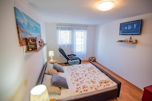 Ein Schlafzimmer mit Bett, Stuhl, Fernseher und einem Fenster mit Vorhängen.