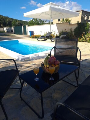 Ein Poolbereich mit Liegestühlen, einem Tisch mit Früchten und Getränken sowie einem weißen Sonnenschirm.