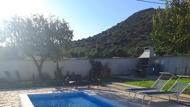 Una piscina rectangular con bordes de piedra, sillas exteriores y vista a una colina.