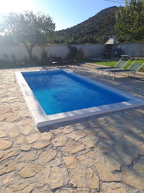 Une piscine rectangulaire entourée de carreaux de pierre, des chaises d’extérieur et une vue sur une colline.