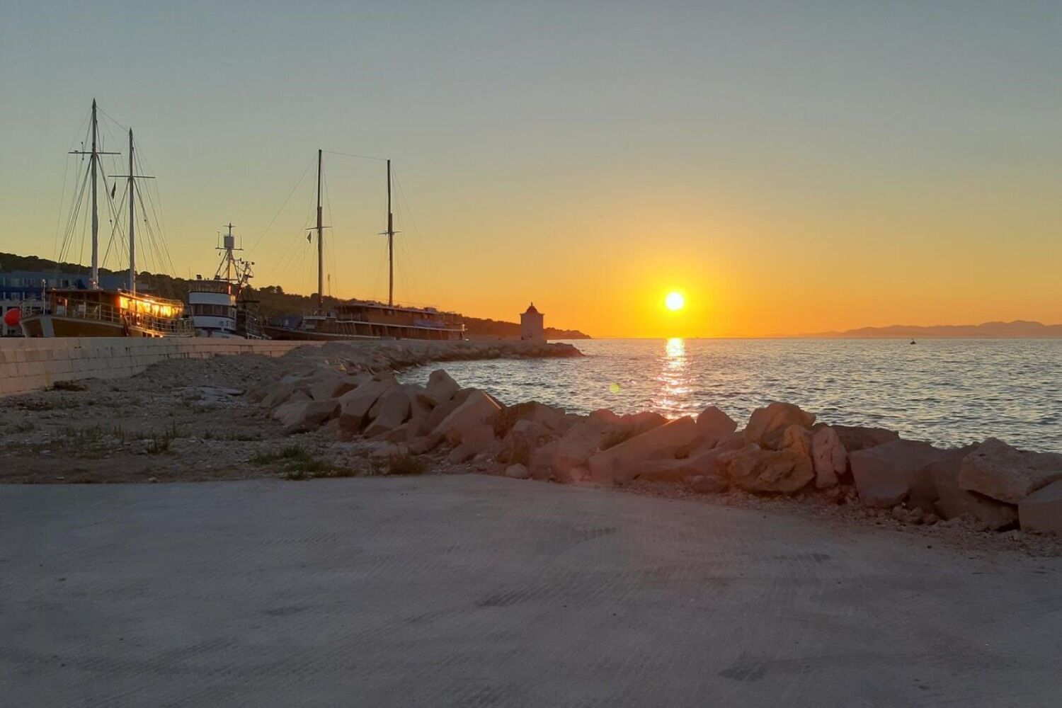A coastal scene with a sunset, rocky shoreline, and a pier with boats.