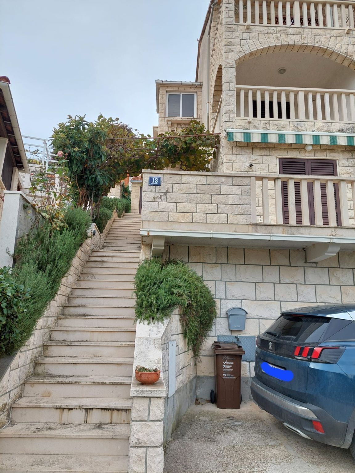 A residential building with a stone facade, a visible number '13' on the wall, a staircase leading to the entrance, and a parked car.