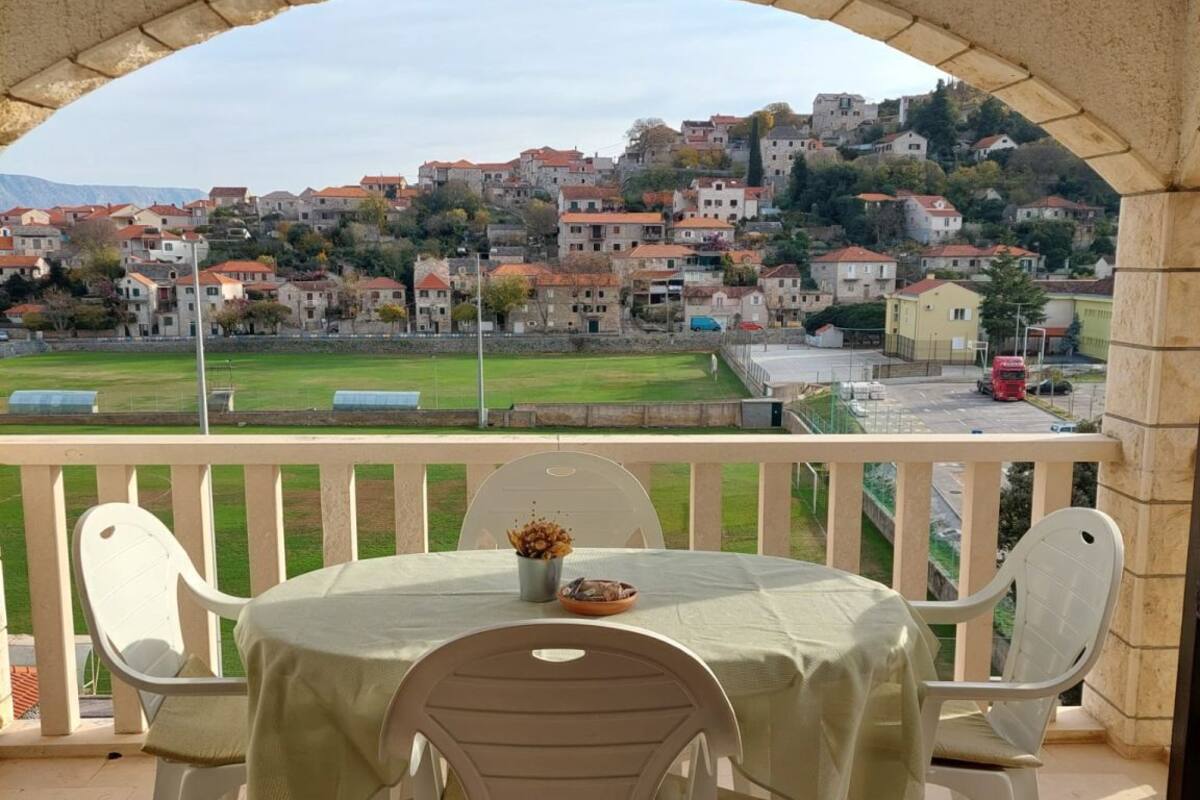 A balcony with a table and chairs, overlooking a town with buildings and a green field.