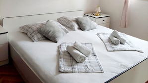 Un lit bien fait, recouvert d’une couverture à carreaux blancs et gris, avec deux serviettes enroulées et une table de chevet surmontée d’une lampe.