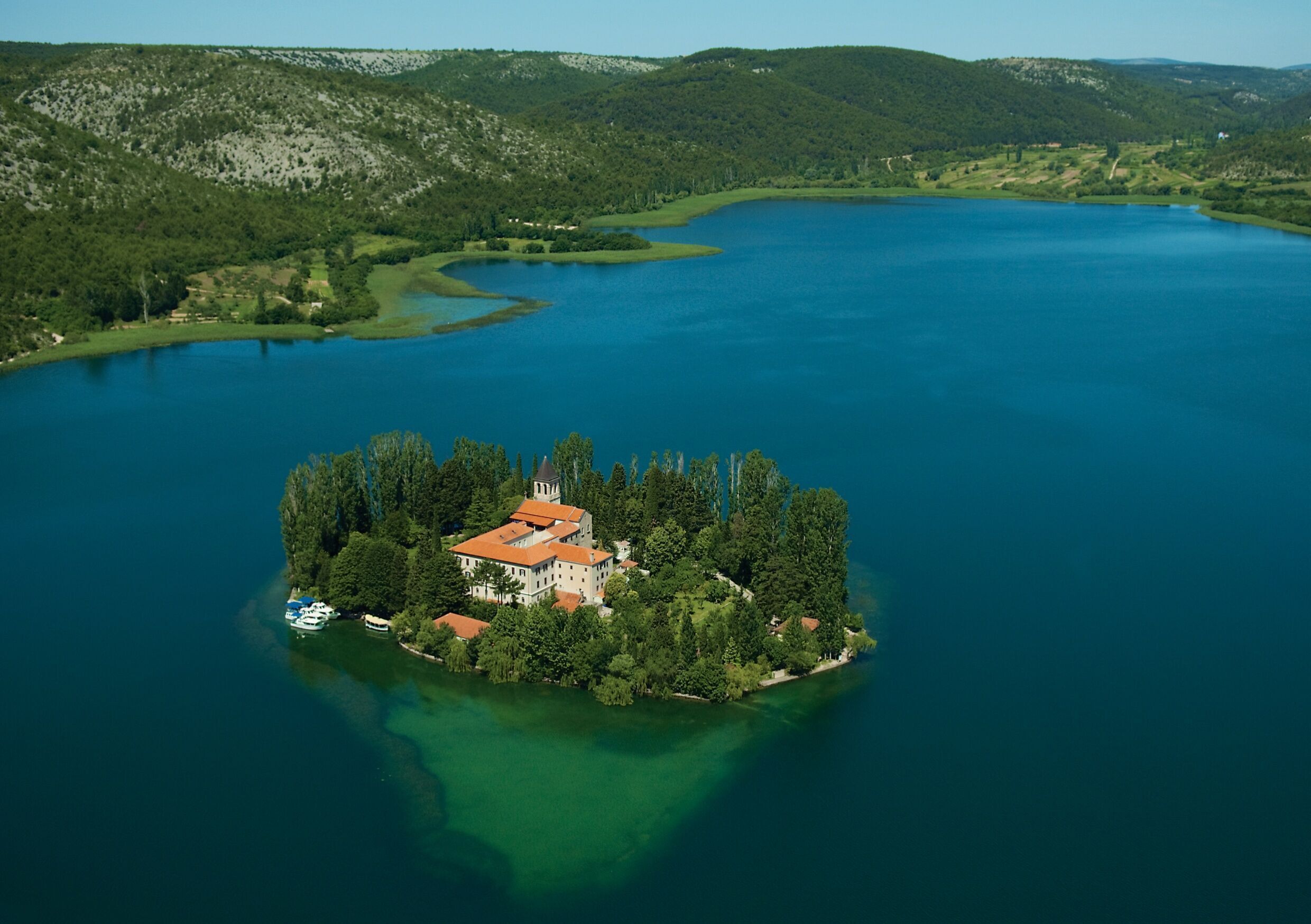 Une vue aérienne d’un lac avec une petite île sur laquelle se trouve un grand bâtiment doté d’un toit rouge, entouré d’une végétation luxuriante et bordé d’une eau d’un bleu limpide.