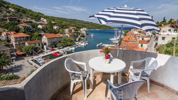 Un balcone con tavolo e sedie, affacciato su una marina con barche e una cittadina costiera.