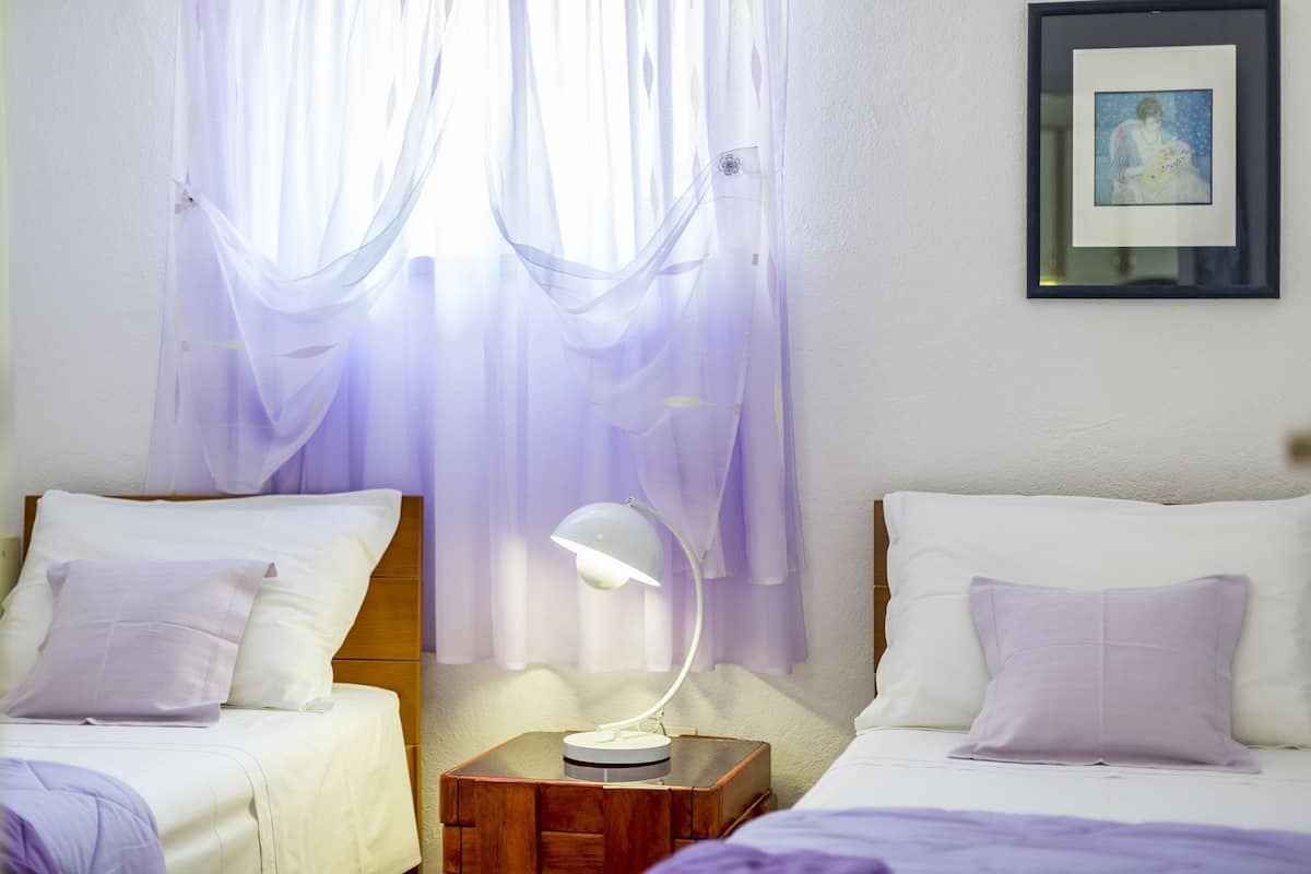 A hotel room with two beds, a bedside table with a lamp, sheer curtains, and a framed picture on the wall.