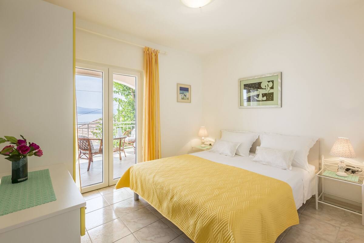 A bedroom with a bed, a nightstand, a lamp, a framed picture, and a view of the outdoors through a sliding door.