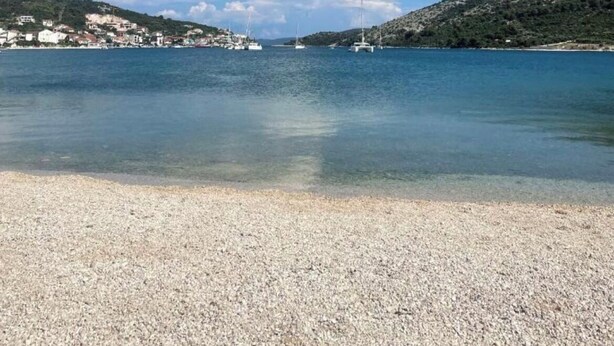 Zona costera con playa de guijarros, agua tranquila y un pueblo lejano con edificios y barcos.