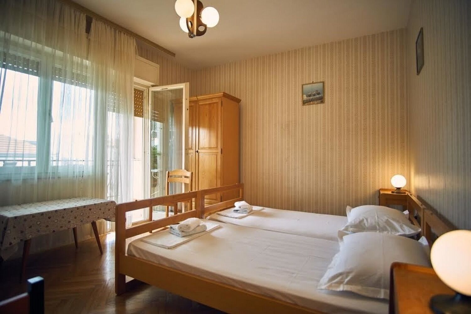 Ein Zimmer mit zwei Einzelbetten, einem Holzschrank, einem Tisch mit Spitzenwäsche und einem Fenster mit durchsichtigen Vorhängen.