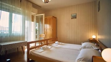 Ein Zimmer mit zwei Einzelbetten, einem Holzschrank, einem Tisch mit Spitzenwäsche und einem Fenster mit durchsichtigen Vorhängen.