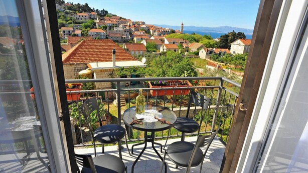 Un balcón con mesa y sillas, con vistas a un pueblo en la ladera con casas de tejados rojos y una vista despejada del mar.