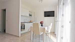 Una cocina moderna con armarios blancos, un microondas y un horno. Hay una zona de comedor con una mesa preparada para dos personas y un televisor montado en la pared.