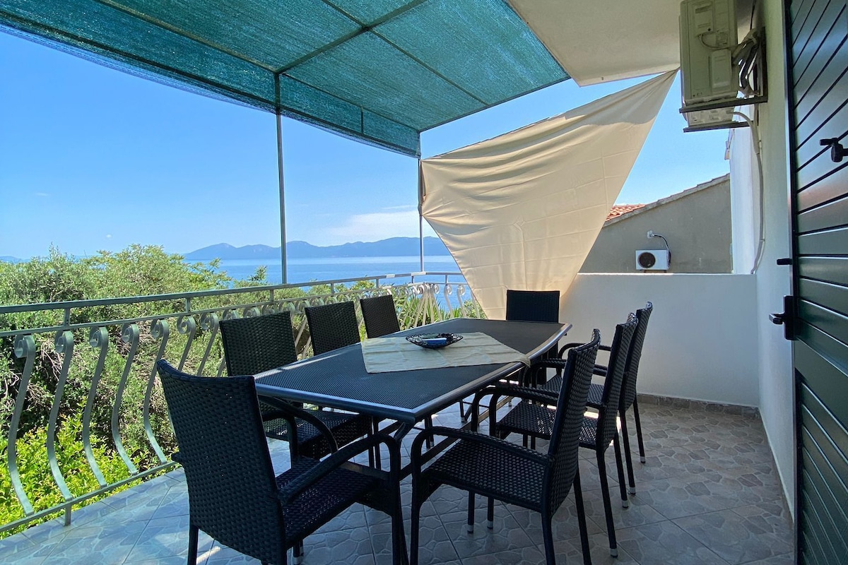 Ein Balkon mit Tisch und Stühlen, Blick auf das Meer und eine weiße Wand mit einem Fenster.