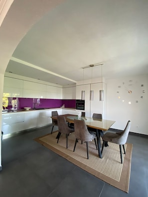 A modern kitchen with white cabinets, a purple backsplash, and a dining area with a glass table and chairs. - Apartments Nada (Sucuraj)