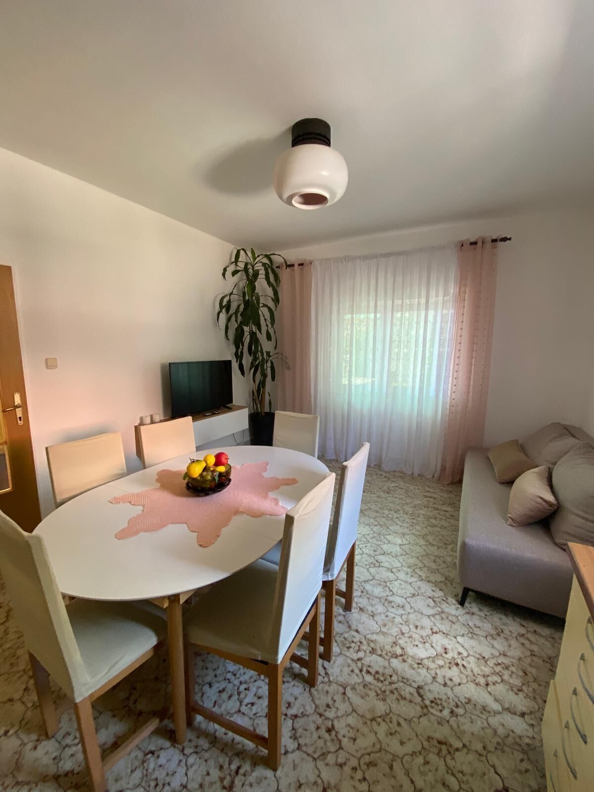 A dining area with a round table, chairs, and a fruit bowl. There is a potted plant, a television, and a sofa in the background.