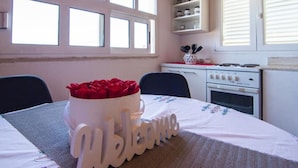A room with a table set for two, a white cabinet with kitchenware, and a window with blinds. - Apartment Ivo (Supetar)