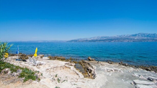 A rocky shoreline with clear blue sea, a few lounge chairs, and a distant mountain range. - Apartment Ivo (Supetar)