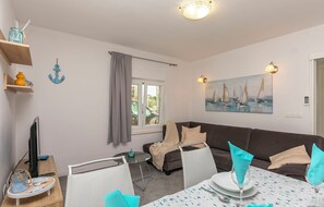 Un salon avec un canapé marron, une table à manger dressée avec des assiettes et des verres, un téléviseur posé sur un meuble et un tableau représentant des voiliers accroché au mur.