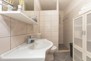 Une salle de bain avec un lavabo, des toilettes et une douche, le tout de couleur blanche. On y trouve des étagères avec des articles de toilette et un porte-serviettes.