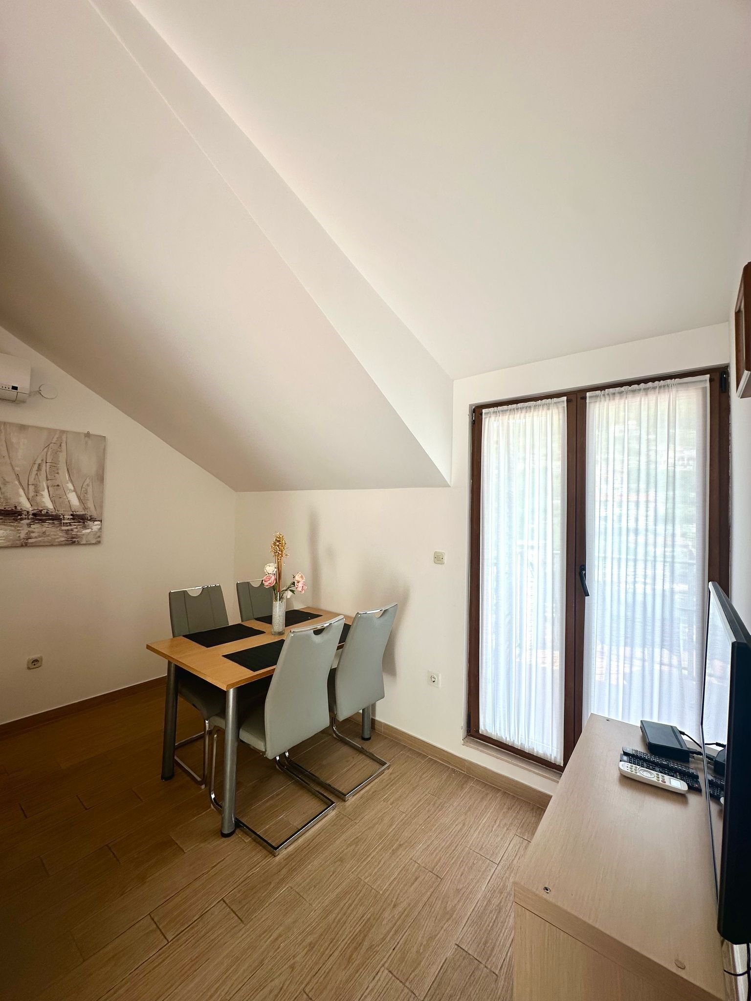 Un comedor con una mesa, sillas y un florero. Se ve una ventana con cortinas y un televisor sobre un mueble.
