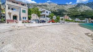 Un bâtiment situé sur la côte, avec une plage de galets devant, une montagne en arrière-plan et des voitures garées le long de la rue.