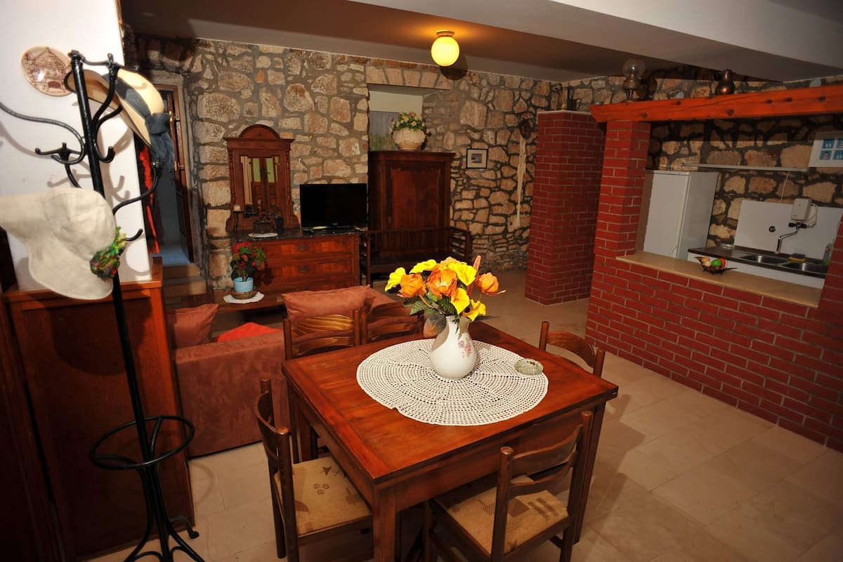 A cozy dining area with a wooden table, chairs, and a vase of yellow flowers. A stone wall and brick fireplace are visible in the background.