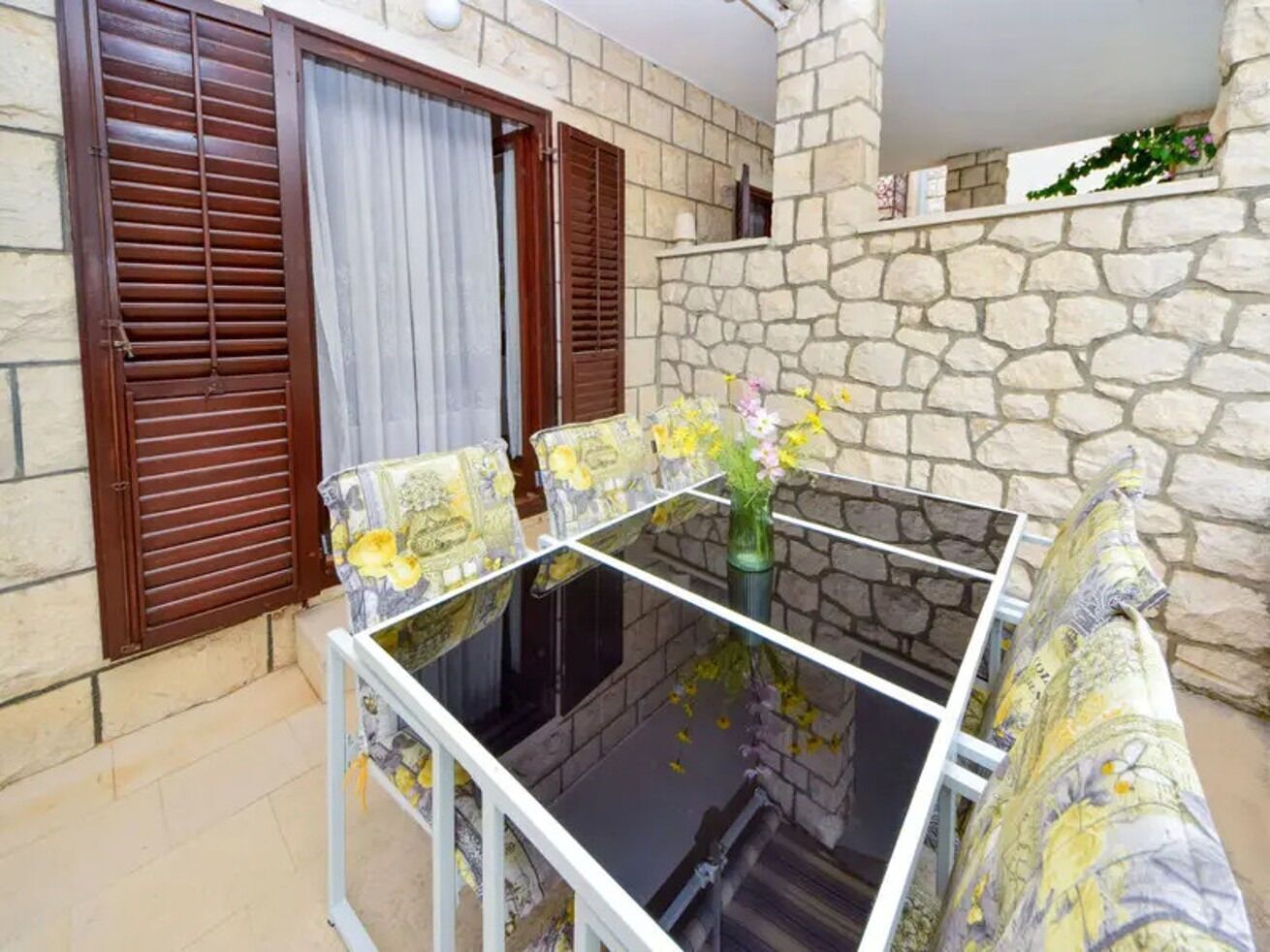 Un balcón con una mesa de vidrio, dos contraventanas de madera y una pared de piedra.