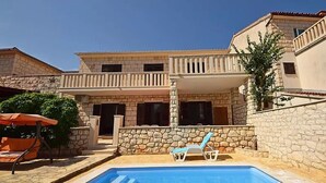 A swimming pool with lounge chairs, a stone wall, and a building with balconies. - Apartment Mari (Supetar)