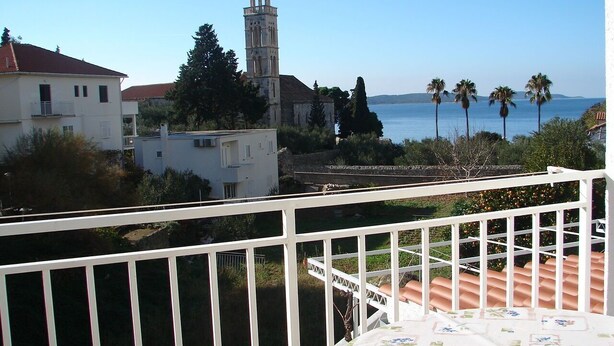 A balcony with a view of a coastal area, including a church tower and palm trees. - Apartment Eta (Hvar)