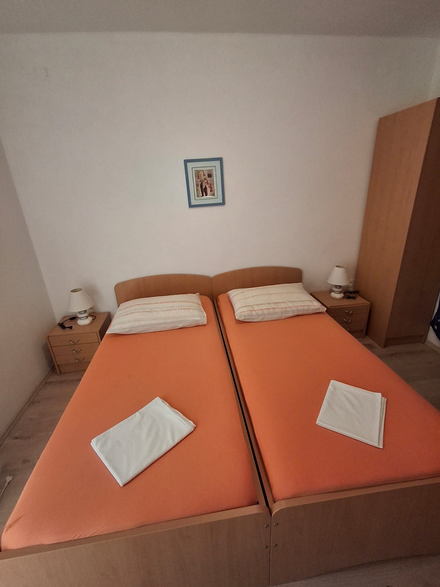 A twin room with two beds, a nightstand with a lamp, a framed picture on the wall, and a wardrobe.