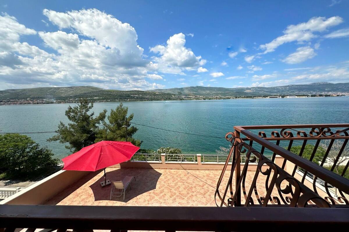 Un balcon avec un parasol rouge, une chaise et une vue sur un lac, avec des montagnes en arrière-plan.