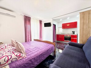 A compact room with a bed, a sofa, a kitchenette with red cabinets, and a TV mounted on the wall. - Apartments Vini (Trogir)