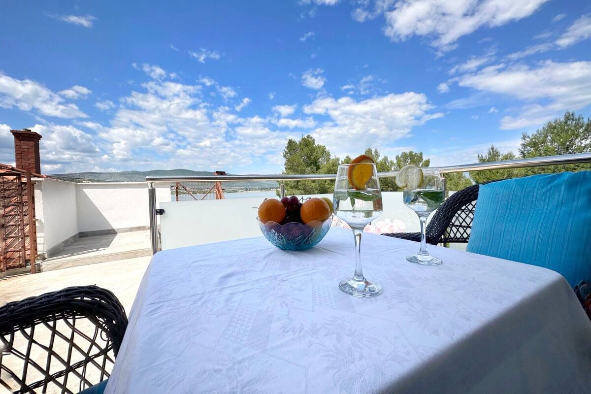 Une table recouverte d’une nappe blanche, un saladier rempli de fruits et deux verres, le tout sur une terrasse avec un ciel dégagé et des collines à l’horizon.