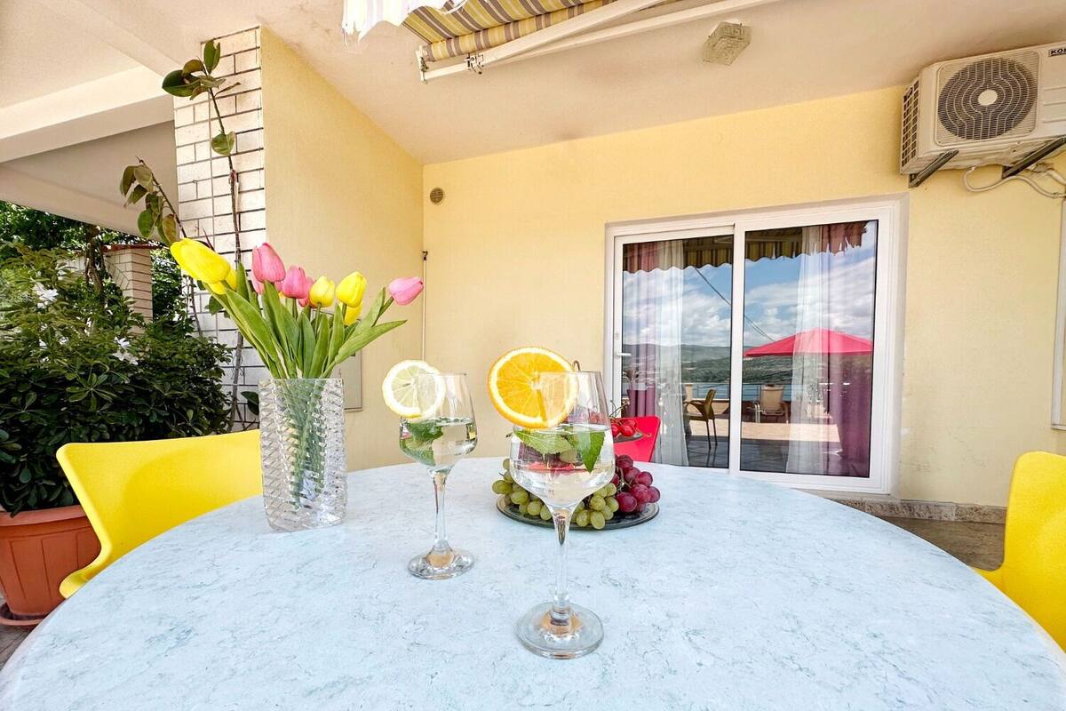 Une table ronde avec un plateau en verre, deux verres à vin, un vase contenant des tulipes jaunes et une garniture de fruits.
