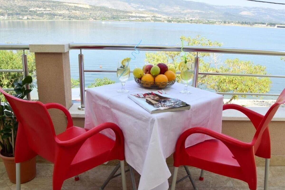 Une table recouverte d’une nappe blanche, des chaises rouges, un saladier rempli de fruits et un magazine, le tout sur un balcon donnant sur un lac.