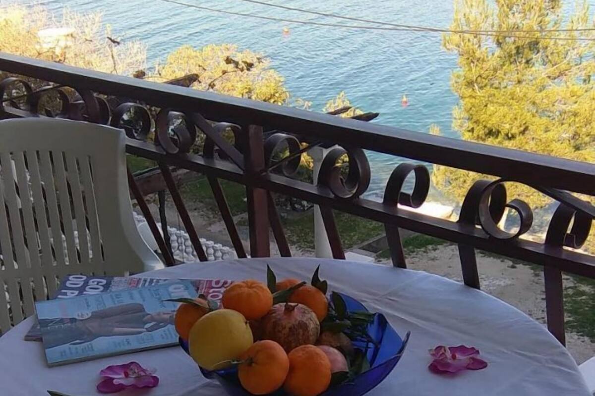 Une table avec un panier de fruits et des fleurs, placée sur un balcon donnant sur une étendue d’eau.