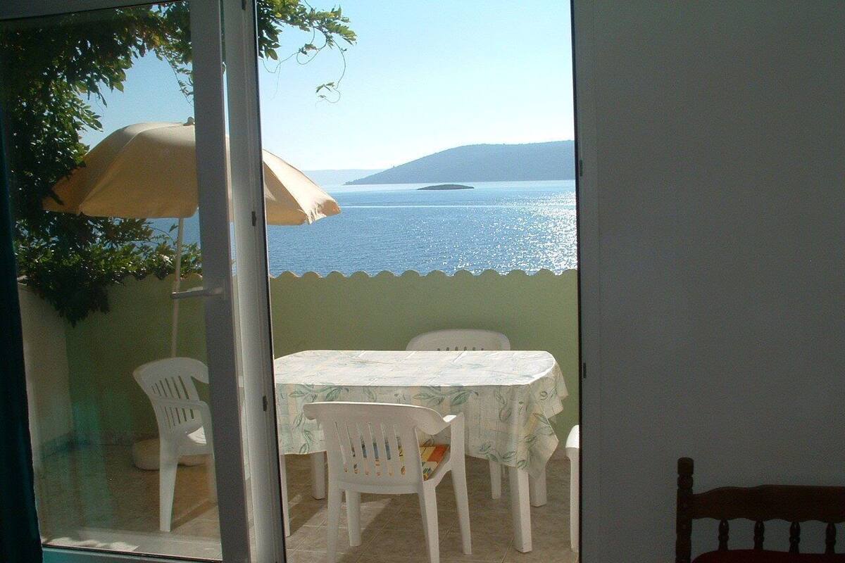 Ein Zimmer mit einer Glasschiebetür, die auf einen Balkon mit Tisch, Stühlen und Sonnenschirm führt, mit Blick aufs Meer.