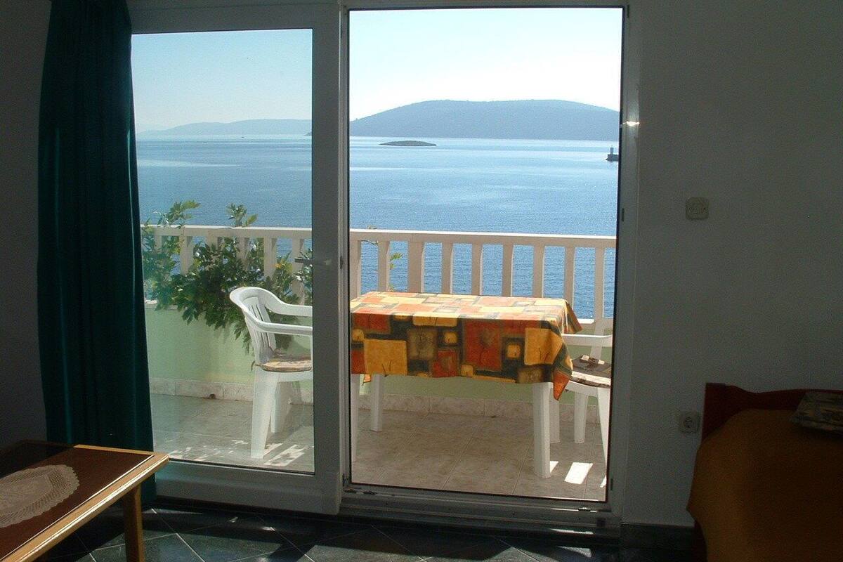Ein Zimmer mit einer Schiebetür, die auf einen Balkon mit Tisch und Stühlen führt, mit Blick auf ein Gewässer und ein entferntes Land.