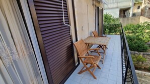 Un balcón con una mesa y sillas, una persiana cerrada y vistas a un jardín y edificios vecinos.