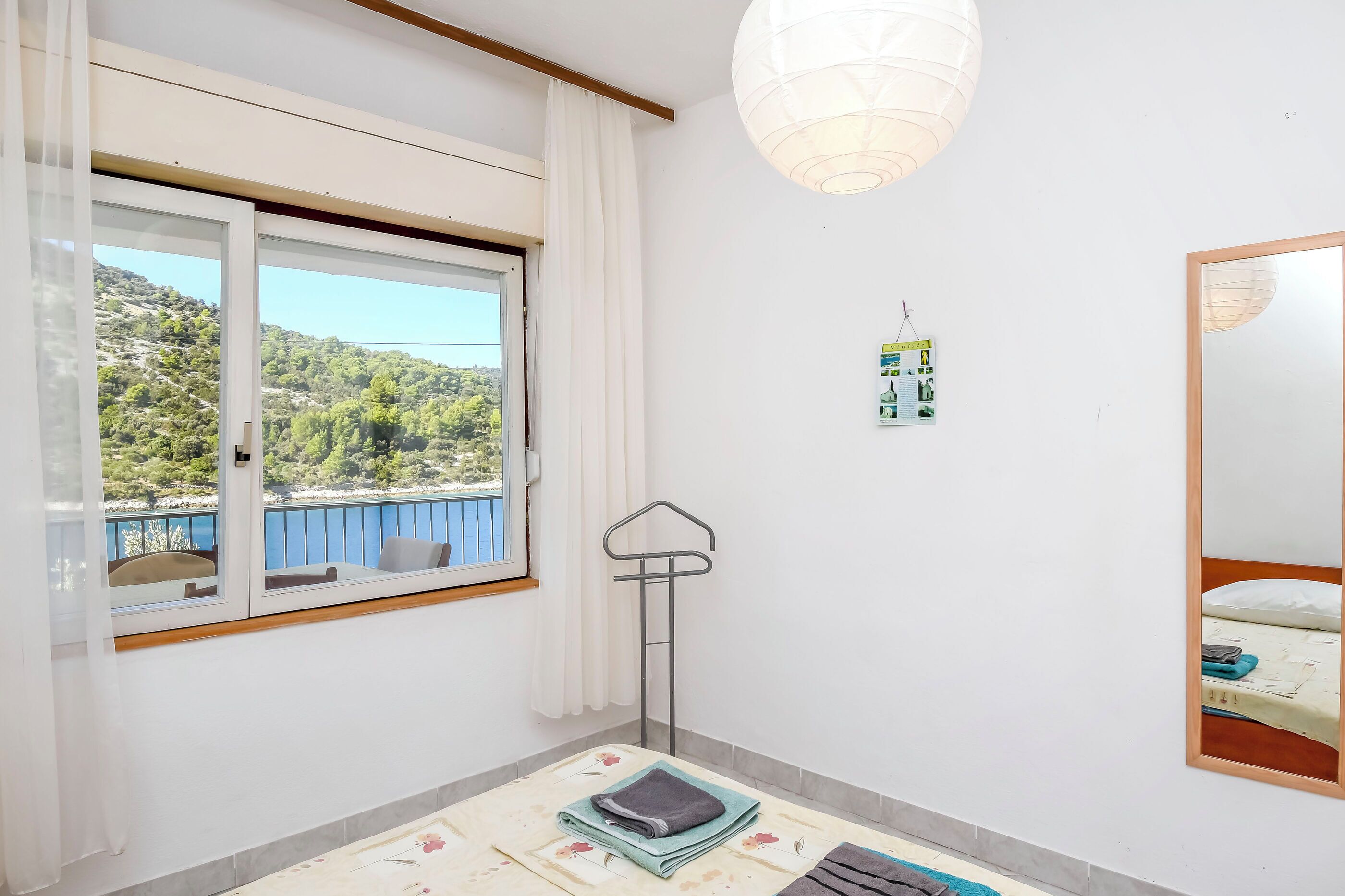 A room with a window overlooking a scenic landscape, a hanging light fixture, a wall-mounted mirror, and a bed with a neatly made bed.
