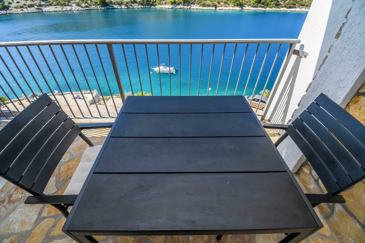 Ein Balkon mit Tisch und zwei Stühlen, mit Blick auf ein türkisblaues Meer und ein Boot.